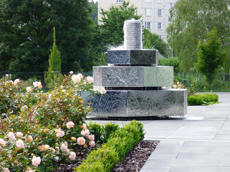 Stupňovitá kamenná fontána s osvětleným válcovým vodotryskem u moderní dlažby a růžových záhonů.