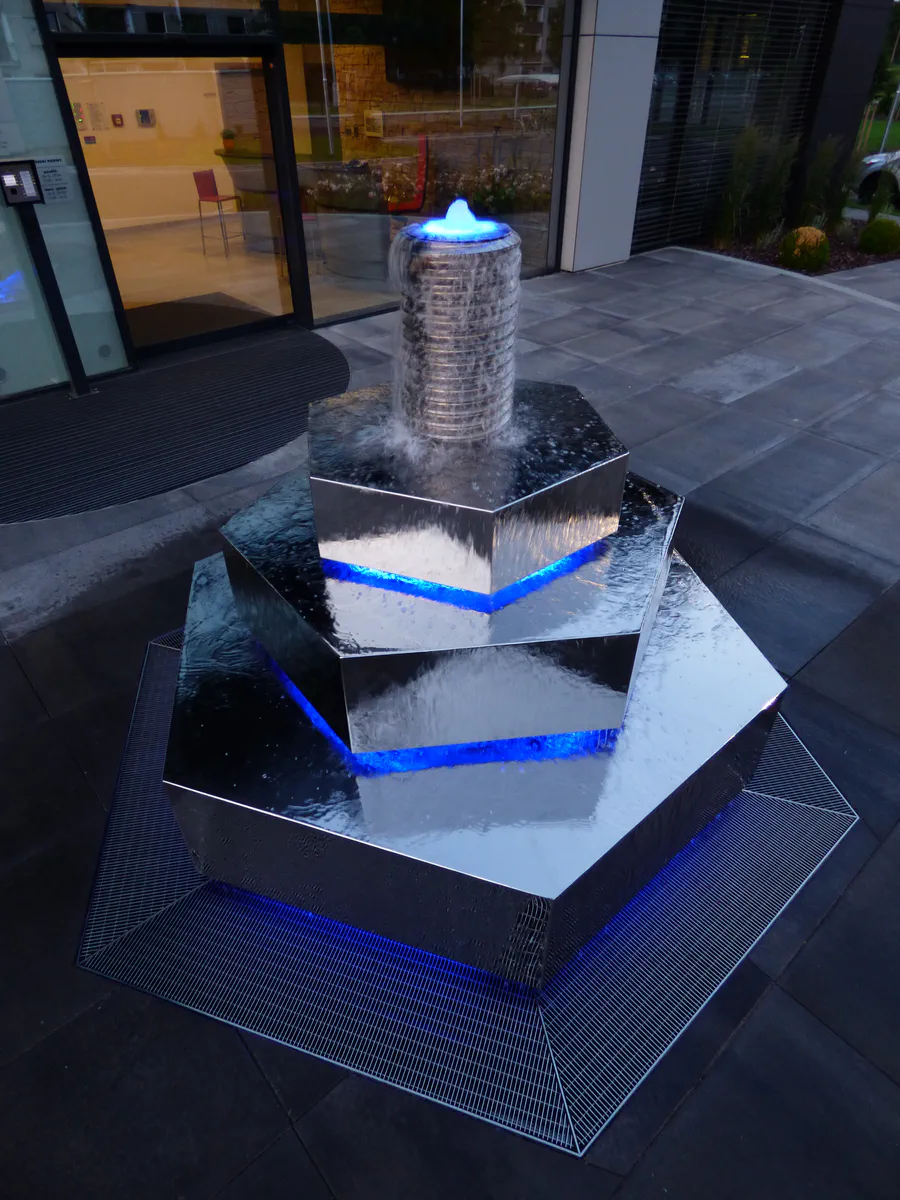 Večerní pohled na fontánu Šroubek s modrým LED osvětlením a vodotryskem před vstupem do budovy.