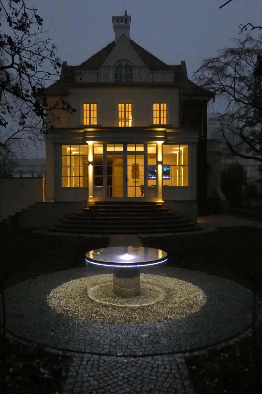 Noční pohled na fontánu před vilou, kde voda stejnoměrně přepadá přes horní misku a vytváří souvislou světelnou clonu.