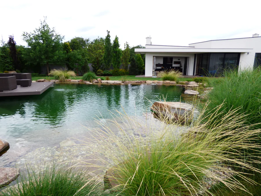 Koupací jezírko s dřevěnou terasou „plovoucí“ nad hladinou, travinami a mělkou kačírkovou zónou před moderní vilou