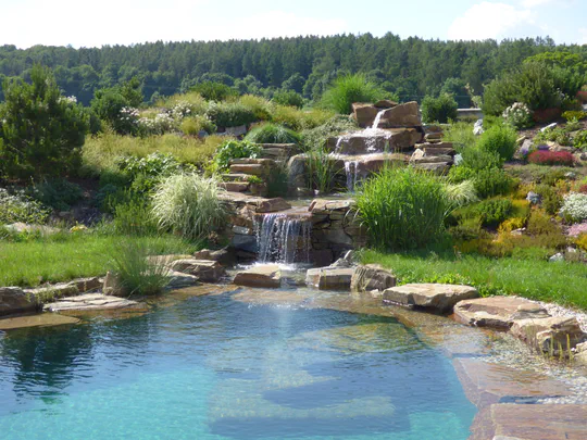 Ukázka vodní kaskády z kamene s lagunou zasazenou do valu osázeného květinami. Laguna je vhodná i ke koupání podobně jako vířivka. Vodní kaskáda z kamene s lagunou zasazená do valu osázeného květinami