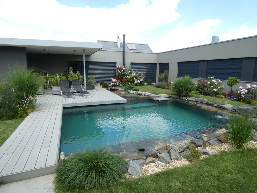 Koupací jezírko s azurovou vodou u terasy moderního domu s růžovými keři v pozadí