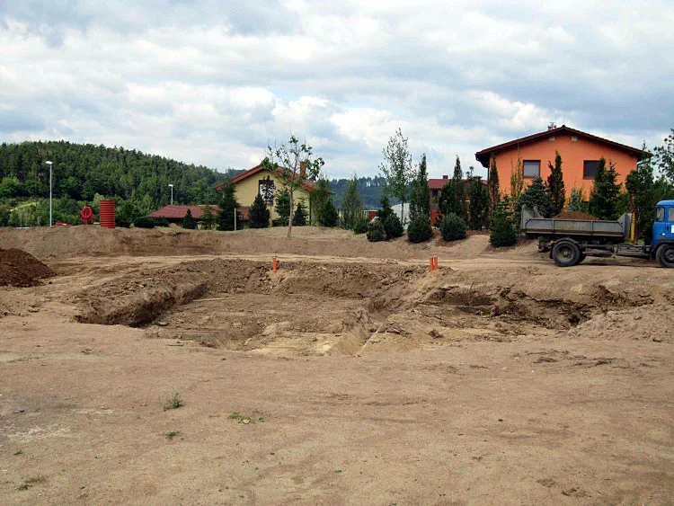 Před realizací Hliněná plocha s vyhloubenou dírou na jezírko veprostřed. Ve vzdálenějším koutu budoucí zahrady jsou vidět čerstvě vysazené dřeviny.