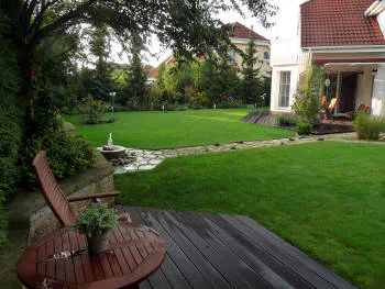 Zahrada s nízce sečeným trávníkem obklopená vzrostlými stromy. Od domu vede kameny dlážděná ceesta k malé fontánce. Na celou scénu je pohled z malé dřevěné terasy se zahradním nábytkem, odkud je fotka pořízena.