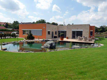 Zatravněná plocha zahrady s jezírkem a domem uprostřed. Jezírko opticky odpovídá velikosti domu a dodává tak celému prostoru dostatečné proporce.